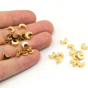 May include: A close-up of a hand holding a pile of gold crescent moon shaped beads. The beads are scattered on a white surface.