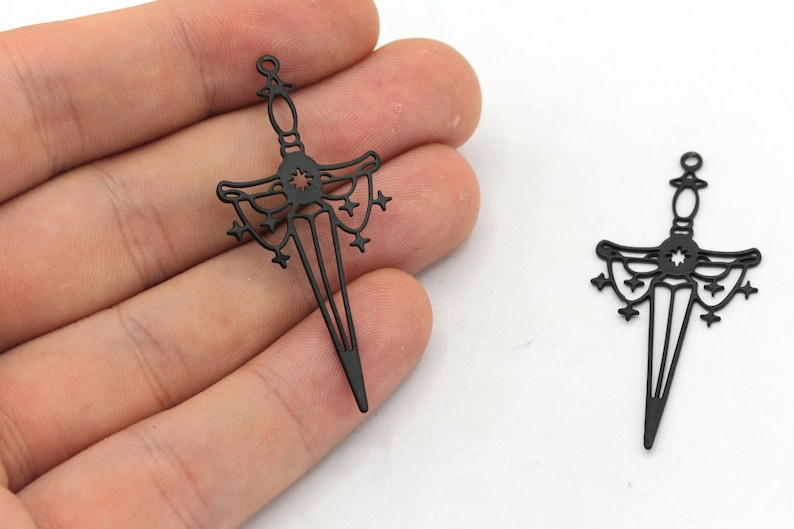 May include: Two black metal sword charms featuring a star design. The swords are detailed with a starburst at the hilt and a decorative design on the blade.