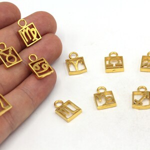May include: Gold-colored metal charms featuring zodiac symbols. Each charm is a small, rectangular shape with a cutout design representing a different astrological sign. The charms have a loop at the top for attaching to jewelry.
