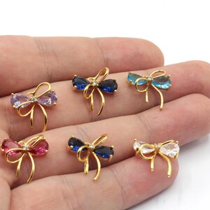 Könnte beinhalten: Sechs goldfarbene, schleifenförmige Schmuckstücke mit farbigen Edelsteinzentren. Die Edelsteine sind in den Farbtönen Rosa, Lila, Blau und Klar. Jede Schleife hat einen kleinen, klaren Akzentstein.