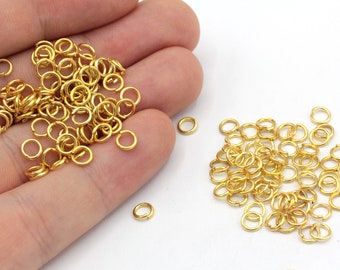 Puede incluir: Un montón grande de anillos de salto dorados, un suministro popular para la fabricación de joyas, se muestra en una mano y en una superficie blanca.
