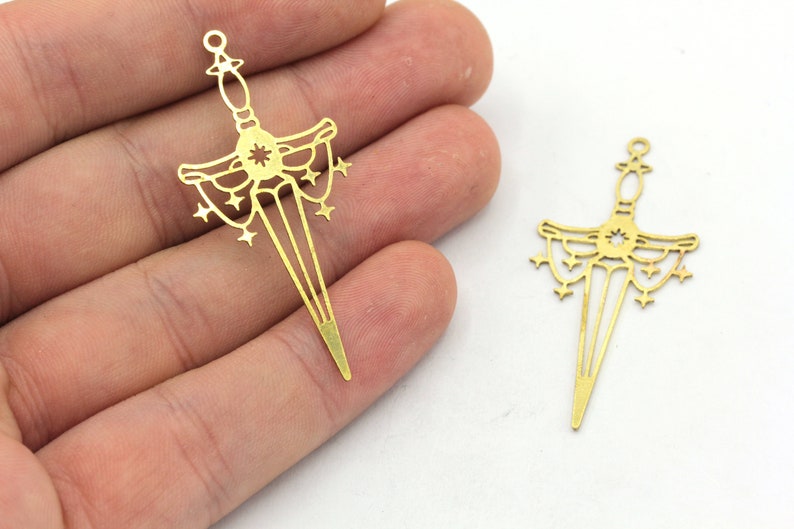 May include: Two gold-toned metal charms in the shape of a sword with a starburst design. The sword has a decorative handle and a pointed tip.