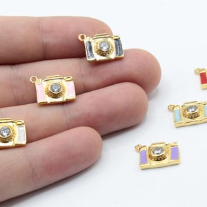 May include: Six gold camera charms with different colored enamel accents. The charms feature a clear rhinestone in the center of each camera lens.