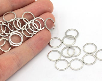 May include: A pile of silver jump rings, approximately 20 in total, are shown on a hand. Another 10 silver jump rings are scattered on a white surface.