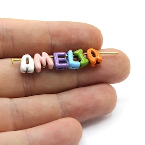 Könnte beinhalten: Ein farbenfroher Perlenname mit der Aufschrift "AMELIA" auf einer goldenen Nadel. Die Perlen sind weiß, rosa, lila, blau, grün und orange. Die Perlen werden von einer Hand gehalten.