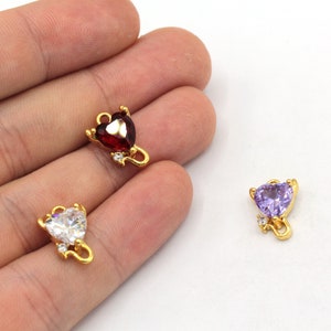May include: Gold-colored heart-shaped charms with a loop for hanging. Each charm features a heart-shaped gemstone in red, white, or purple, accented with a small, clear gemstone. These charms are likely for jewelry making.