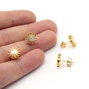 May include: A pair of gold earrings with a white stone in the center of each earring. The earrings are shaped like a sun with rays extending from the center.