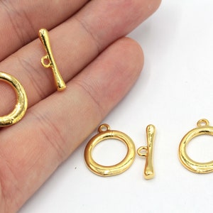 May include: Several gold-colored toggle clasps. The clasps consist of a circular ring and a bar with a loop. The clasps are likely for jewelry making or other craft projects. The clasps are shown against a white background.