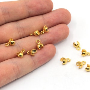 Puede incluir: Un conjunto de pequeños hallazgos de joyería dorados, probablemente tapas de cuentas, dispersos sobre una superficie blanca. Cada pieza tiene un cuerpo texturizado y redondeado con un diseño dividido en forma de pétalo en la parte superior y un pequeño lazo. Estos hallazgos se utilizan en la fabricación de joyas.