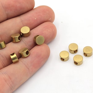 May include: A collection of small, round, brass-colored beads with a hole through the center. The beads are cylindrical with a flat top and bottom. The beads are shown against a white background.