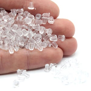 May include: A pile of clear plastic earring backs in a hand. The backs are small and round with a small hole in the center.