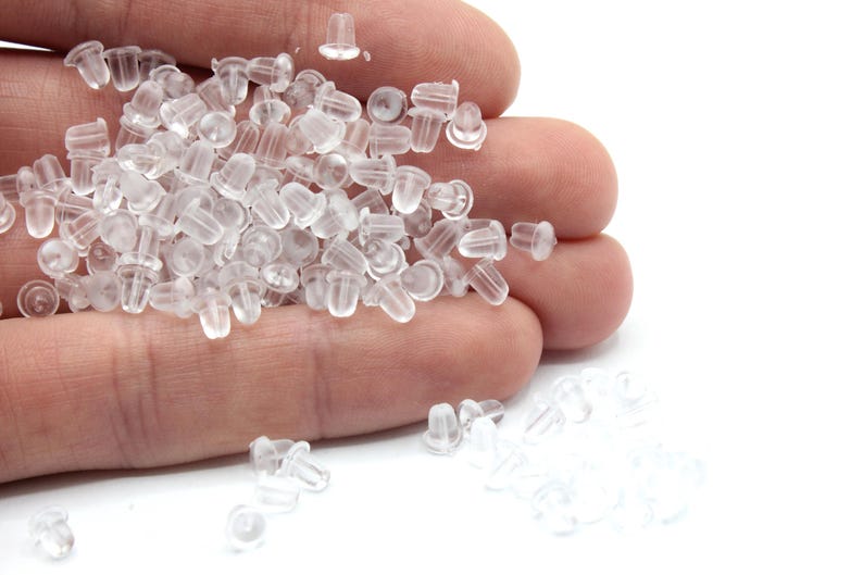 May include: A pile of clear plastic earring backs in a hand. The backs are small and round with a small hole in the center.