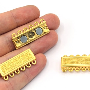 Pode incluir: Dois fechos magnéticos dourados para colares ou pulseiras. Cada fecho tem cinco laços e um design retangular texturizado. Um fecho tem dois ímãs visíveis.