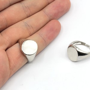 May include: A silver signet ring with a smooth, oval face is shown on a hand, with another ring next to it. The rings have a polished finish and a classic design, suitable for everyday wear or special occasions.