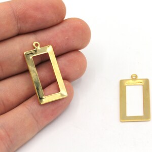 May include: Two gold-colored rectangular pendant charms with a hollow center and a small loop at the top for attaching to a chain or other jewelry component. The charms are shown against a white background.
