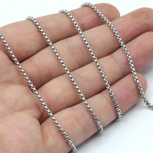 May include: A silver chain necklace with a delicate, box chain design. The chain is shown laid out on a hand, demonstrating its length and flexibility.