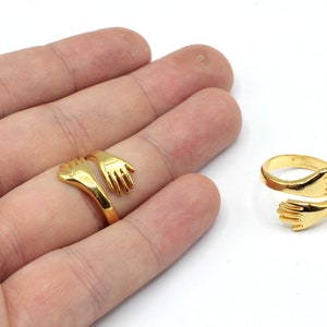 May include: A gold-toned ring featuring two hands clasped together in a hug. The ring is open and adjustable to fit different finger sizes.