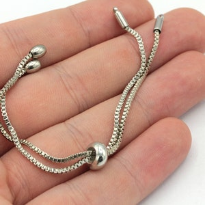 May include: A silver chain bracelet with a sliding bead closure. The bracelet is made of two thin chains that are connected by a small bead.
