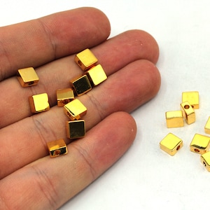 May include: A collection of small, shiny, gold-colored square beads. Each bead has a hole running through the center, suggesting they are designed for stringing. The beads are displayed against a white background.