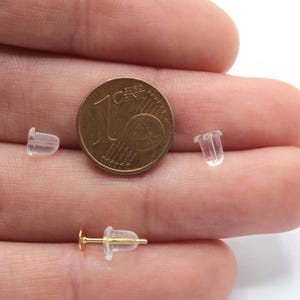May include: Close-up of three clear plastic earring backs next to a 1 Euro coin. The earring backs are small and translucent, and the coin is a standard size.