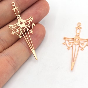 May include: Two gold-coloured metal sword charms with a starburst design and a decorative border. The charms are shown against a white background.