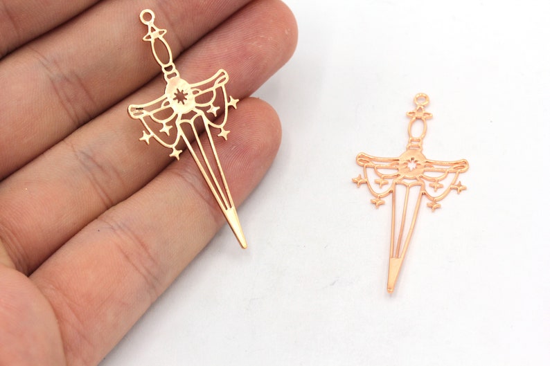 May include: Two gold-coloured metal sword charms with a starburst design and a decorative border. The charms are shown against a white background.