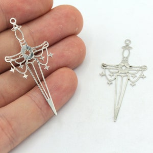 May include: Two silver-toned metal sword charms with a starburst design. The swords have a pointed tip and a decorative handle with a starburst design.