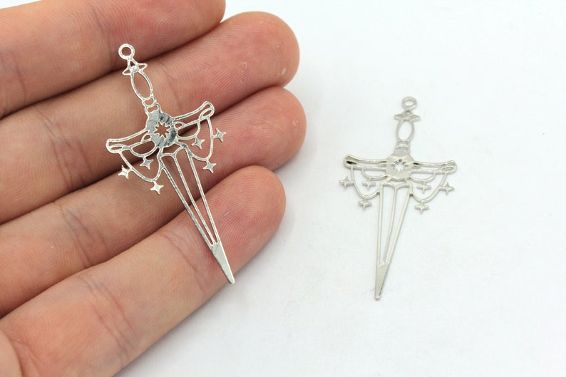 May include: Two silver-toned metal sword charms with a starburst design. The swords have a pointed tip and a decorative handle with a starburst design.