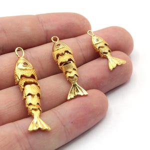 Può includere: Tre ciondoli a forma di pesce dorati con un piccolo occhio di pietra preziosa rossa. I ciondoli sono appesi a un anello nella parte superiore e hanno un design dettagliato e testurizzato.