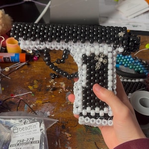 May include: A handmade beaded pistol in black and white, with a decorative silver charm. The gun is constructed from small beads, with a dark purple barrel. The image also shows a bag of beads with the text "beadtin".