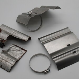 Puede incluir: Cuatro escudos t&eacute;rmicos de metal para un sistema de escape. Dos est&aacute;n intactos, uno est&aacute; parcialmente da&ntilde;ado y uno est&aacute; en pedazos. Los escudos son plateados y tienen una superficie corrugada. Hay dos abrazaderas de metal plateadas.