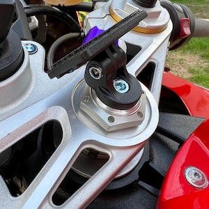 Może przedstawiać: Zbliżenie przedniego zawieszenia i kierownicy motocykla. Czarny uchwyt kamery jest przymocowany do srebrnych metalowych elementów. Motocykl ma czerwone i czarne akcenty, z widocznym fioletowym gumowym elementem.