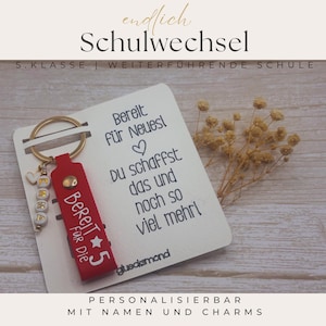 Könnte beinhalten: Ein roter Schlüsselanhänger aus Leder mit goldenem Ring, bedruckt mit 'Bereit für die 5' und einem Stern. Er enthält einen weißen Perlenanhänger mit dem Namen 'EMMA' und einem Herz. Der Anhänger ist auf einer weißen Karte mit dem Text 'Bereit für Neues' befestigt.