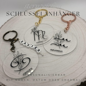 Könnte beinhalten: Transparente Acryl-Schlüsselanhänger mit Tierkreiszeichen und floralen Designs. Die Schlüsselanhänger zeigen die Wörter "VIRGO", "AQUARIUS" und "CANCER" in Schwarz. Ein Schlüsselanhänger hat den Namen "XAVIER" in weißen Perlen. Der Text "SCHLÜSSELANHÄNGER" steht oben.