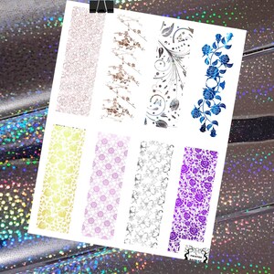 May include: A sheet of nail art decals featuring various floral and abstract designs. The decals include patterns of roses, branches, and swirls in colors like rose gold, brown, silver, blue, yellow, pink, and purple. The sheet is held by a black binder clip.