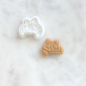 May include: A white plastic cookie cutter and a baked cookie shaped like a croissant with a bow on top. The cookie is light brown with detailed lines, and the cutter is a simple outline.