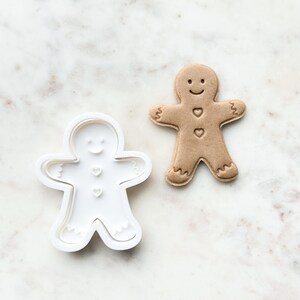 May include: A white plastic gingerbread man cookie cutter and a brown gingerbread man cookie. Both have a smiling face and three heart-shaped buttons.