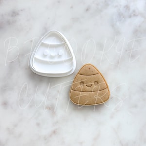Peut inclure: Un emporte-pièce en plastique blanc en forme de bonbon de maïs à côté d'un biscuit en forme de bonbon de maïs cuit. L'emporte-pièce et le biscuit ont tous deux la forme d'un bonbon de maïs avec des lignes horizontales et un visage souriant.