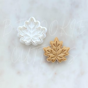 May include: A white plastic maple leaf cookie cutter and a matching light brown maple leaf shaped cookie. The cutter features a detailed interior vein design that imprints onto the dough, creating a crisp leaf shape with defined edges.