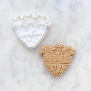 May include: A white plastic cookie cutter shaped like a cluster of three carrots with leafy tops, alongside a light brown cookie dough cutout featuring the same carrot design with embossed details including horizontal lines and a small bow.