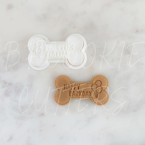 May include: A bone-shaped cookie cutter and a baked cookie. The cookie cutter is white and has the words "HAPPY BARKDAY" and a paw print design. The baked cookie is light brown and has the same design.