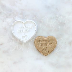 May include: A white heart-shaped cookie cutter and a matching tan heart-shaped biscuit. Both feature the embossed text ALWAYS + FOREVER and a graphic of two interlocking rings. The cutter is made of white plastic and the biscuit is baked dough.