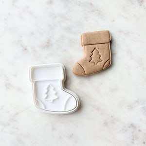 May include: A white plastic cookie cutter shaped like a Christmas stocking with a small Christmas tree design inside. Next to it is a brown cookie in the shape of a stocking with a Christmas tree design.
