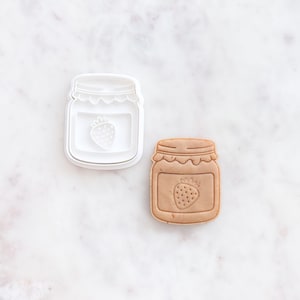 May include: A white plastic cookie cutter shaped like a jar of strawberry jam, alongside a baked cookie in the same form. The cutter features a strawberry design. The baked cookie is light brown in colour.