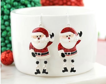 Puede incluir: Un par de pendientes colgantes de plata con un diseño de Papá Noel rojo y blanco con botas negras y barba blanca. Las figuras de Papá Noel sostienen una perla blanca en su mano derecha y saludan con la izquierda.