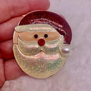 Puede incluir: Un pin de Santa Claus brillante, rojo y blanco con un acento de perla. Santa Claus lleva un gorro rojo y tiene barba y bigote blancos.