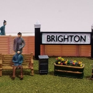 Può includere: Una scena di stazione ferroviaria in miniatura con un cartello che recita "BRIGHTON" e diverse figure in miniatura, tra cui una panchina, fioriere e cassette postali.
