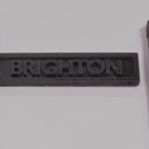 May include: Black plastic sign with the word "BRIGHTON" in raised letters, flanked by two black plastic posts with pointed tops.