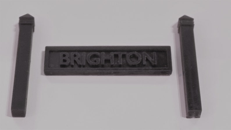 May include: Black plastic sign with the word "BRIGHTON" in raised letters, flanked by two black plastic posts with pointed tops.