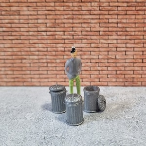 Puede incluir: Cuatro cubos de basura de metal gris con tapas, dispuestos en círculo sobre una superficie de hormigón gris. Una persona en miniatura se encuentra en el centro del círculo.